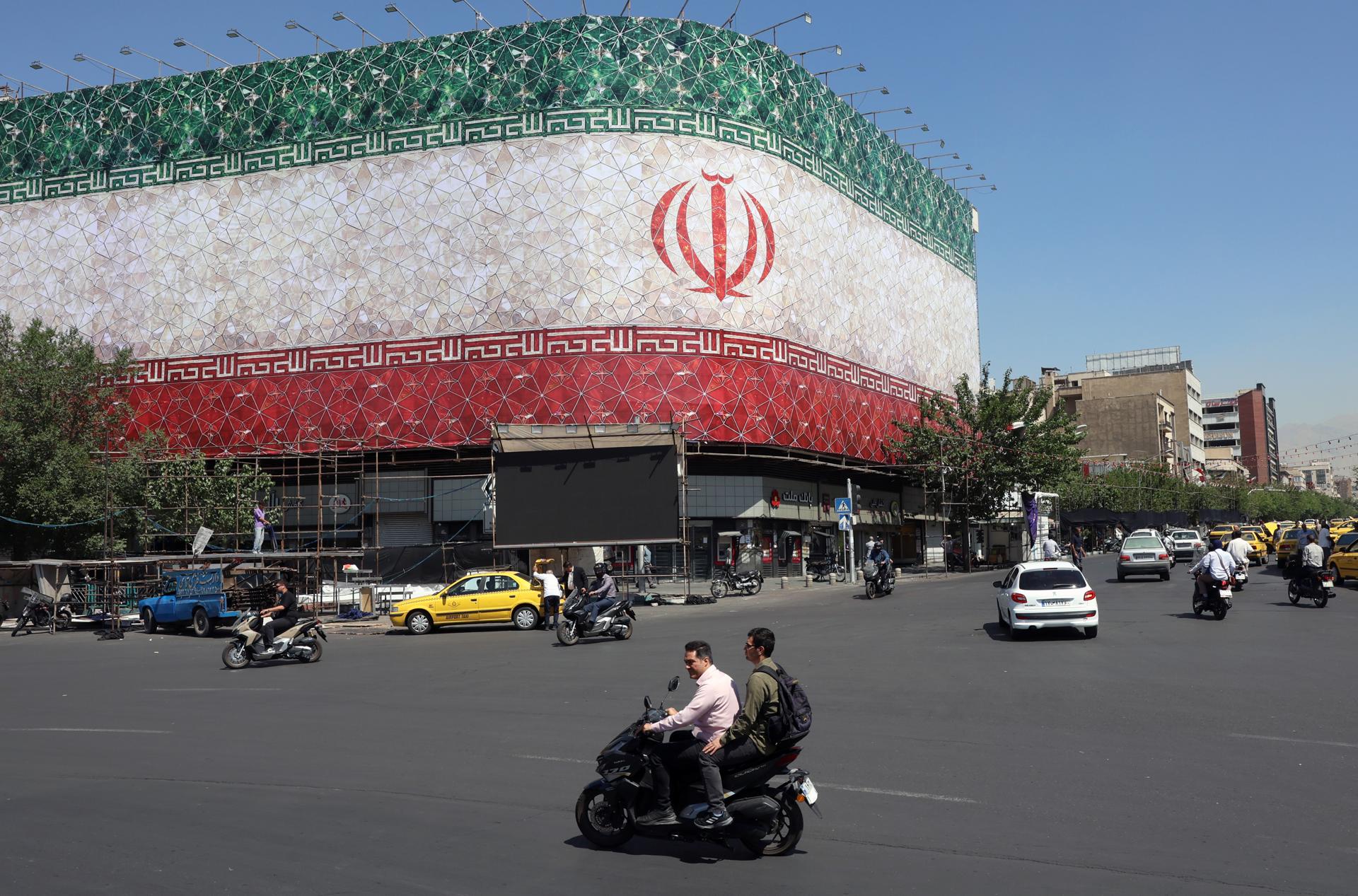 Unos iraníes pasan en coche junto a una valla publicitaria con la bandera nacional de Irán en la plaza Enqelab de Teherán, Irán. EFE/Abedin Taherkenareh